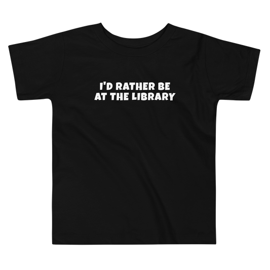 "I’d rather be at the library" printed on a black toddler short sleeve tee laying on a flat surface
