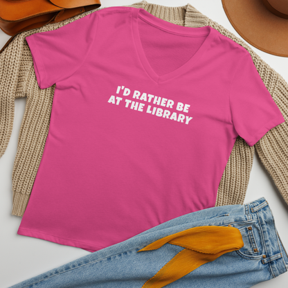 "I’d rather be at the library" printed on a berry women's relaxed v-neck t-shirt laying on a flat surface amongst other clothes