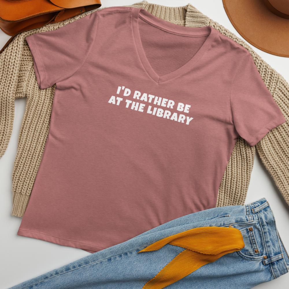 "I’d rather be at the library" printed on a heather mauve women's relaxed v-neck t-shirt laying on a flat surface amongst other clothes