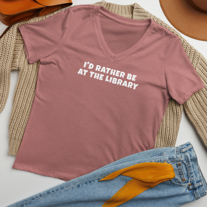 "I’d rather be at the library" printed on a heather mauve women's relaxed v-neck t-shirt laying on a flat surface amongst other clothes
