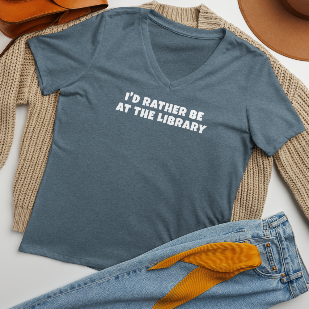 "I’d rather be at the library" printed on a heather slate women's relaxed v-neck t-shirt laying on a flat surface amongst other clothes