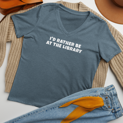"I’d rather be at the library" printed on a heather slate women's relaxed v-neck t-shirt laying on a flat surface amongst other clothes