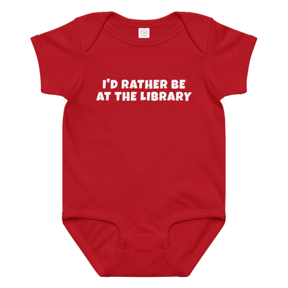 "I’d rather be at the library" printed on a red baby jersey bodysuit laying on a flat surface amongst other clothes