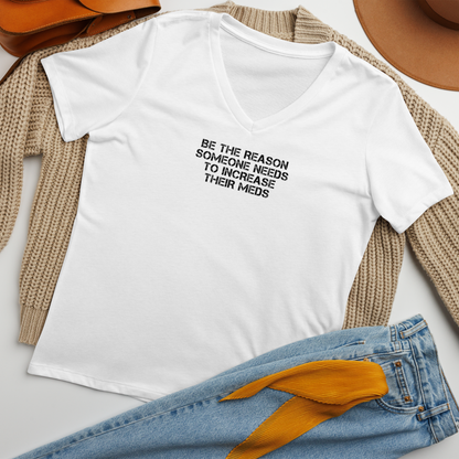 "Be the reason someone needs to increase their meds" printed on a solid white blend women's relaxed v-neck t-shirt laying on a flat surface amongst other clothes