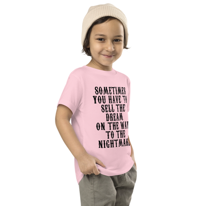 "Sometimes you have to sell the dream on the way to the nightmare" printed on a pink toddler short sleeve tee worn by a male who is also wearing a knit cap