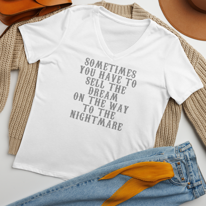 "Sometimes you have to sell the dream on the way to the nightmare" printed on a solid white blend women's relaxed v-neck t-shirt laying on a flat surface amongst other clothes