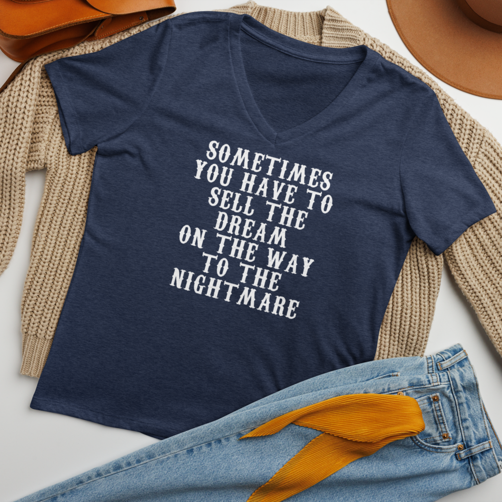 "Sometimes you have to sell the dream on the way to the nightmare" printed on a heather navy women's relaxed v-neck t-shirt laying on a flat surface amongst other clothes