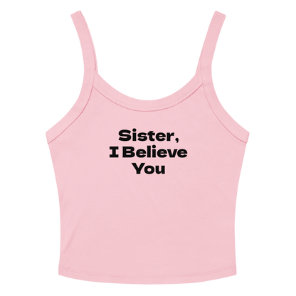 "Sister, I Believe You!" printed on a solid pink blend women's micro-rib tanktop laying on a flat surface amongst sunglasses and shorts