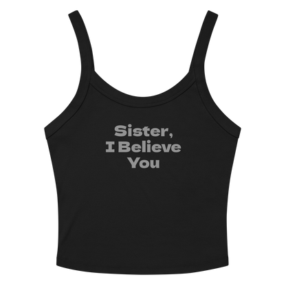 "Sister, I Believe You!" printed on a solid black blend women's micro-rib tanktop laying on a flat surface amongst sunglasses and shorts