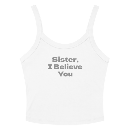 "Sister, I Believe You!" printed on a solid white blend women's micro-rib tanktop laying on a flat surface amongst sunglasses and shorts