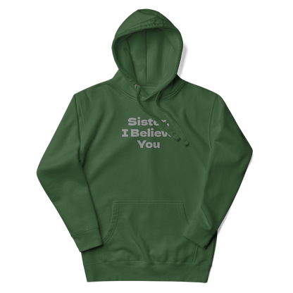 "Sister, I Believe You!" printed on a forest green unisex hoodie laying on a flat surface with the hood up