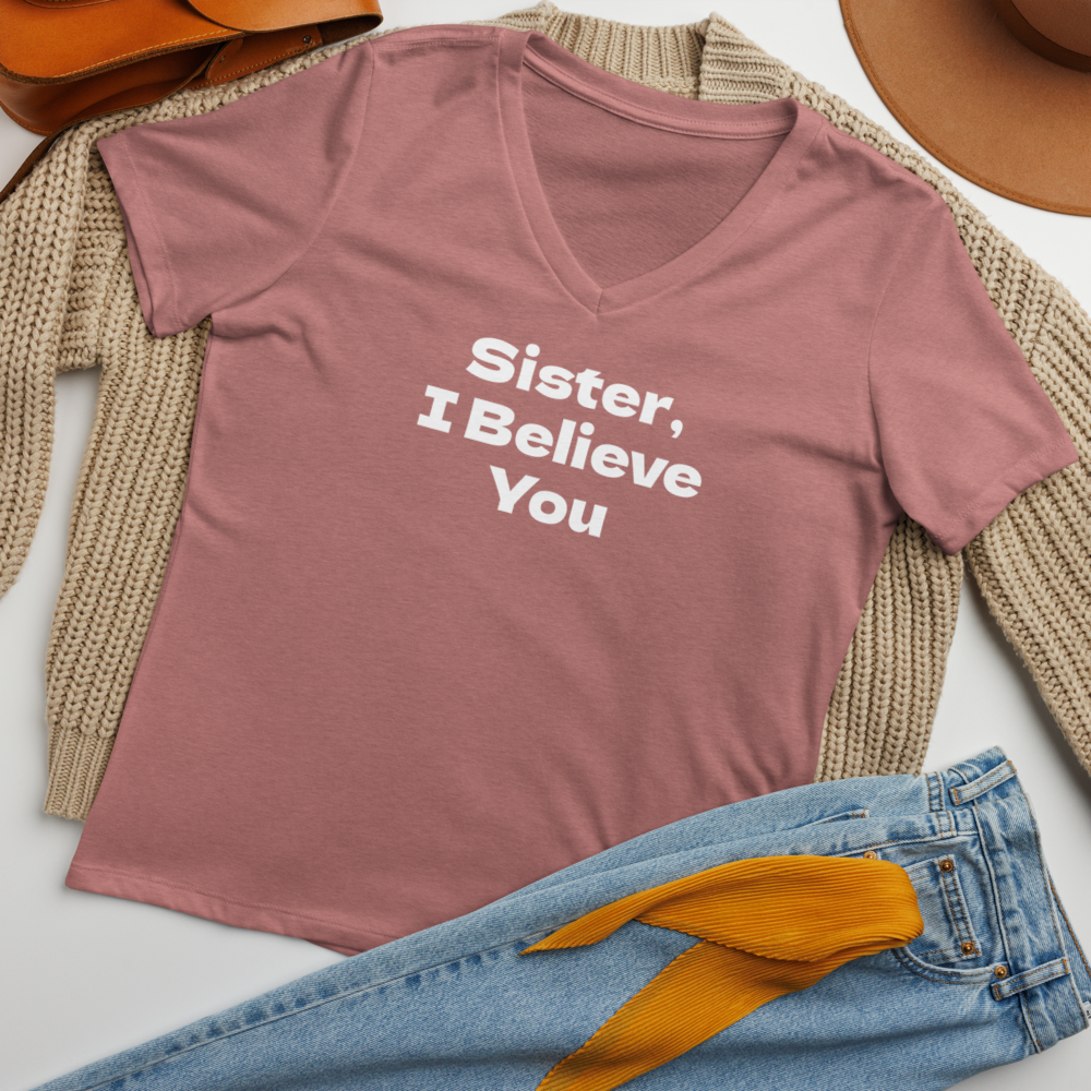 "Sister, I Believe You!" printed on a heather mauve women's relaxed v-neck t-shirt laying on a flat surface amongst other clothes