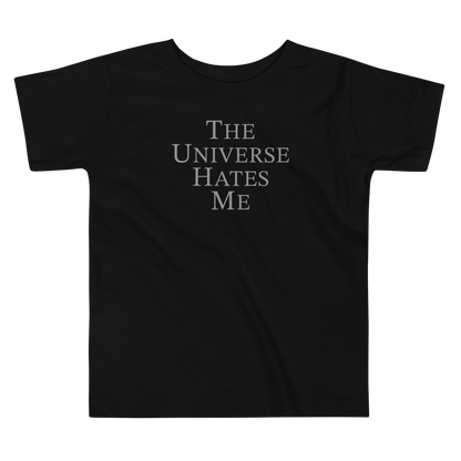 "The Universe Hates Me" printed on a black toddler short sleeve tee laying on a flat surface