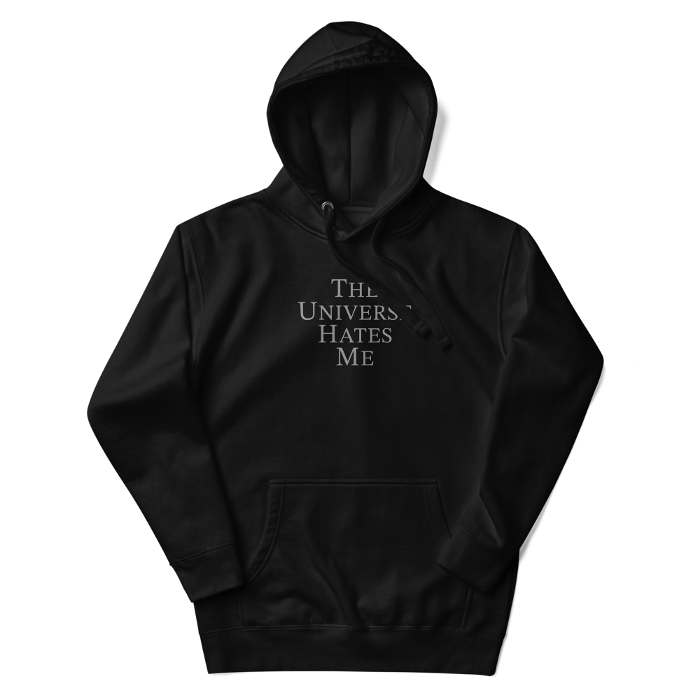 "The Universe Hates Me" printed on a black unisex hoodie laying on a flat surface with the hood up