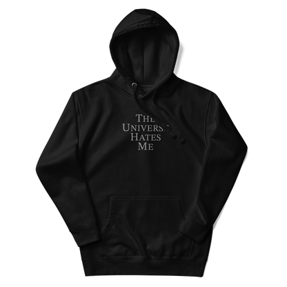 "The Universe Hates Me" printed on a black unisex hoodie laying on a flat surface with the hood up