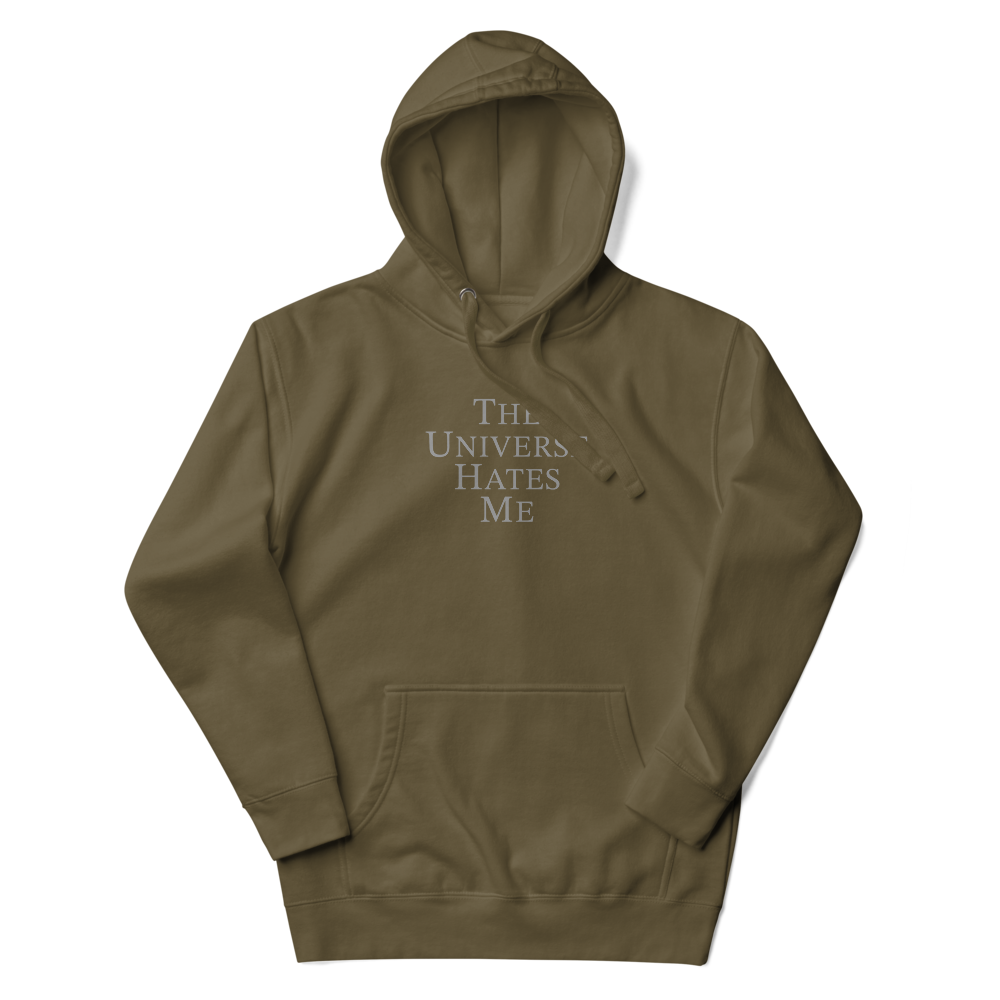 "The Universe Hates Me" printed on a military green unisex hoodie laying on a flat surface with the hood up