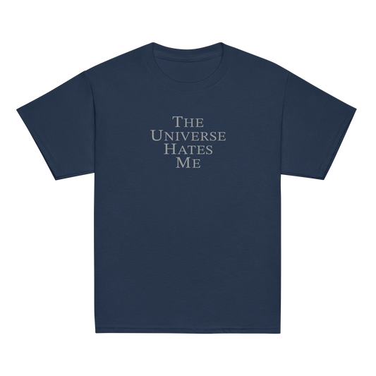 "The Universe Hates Me" printed on a navy youth classic tee laying on a flat surface