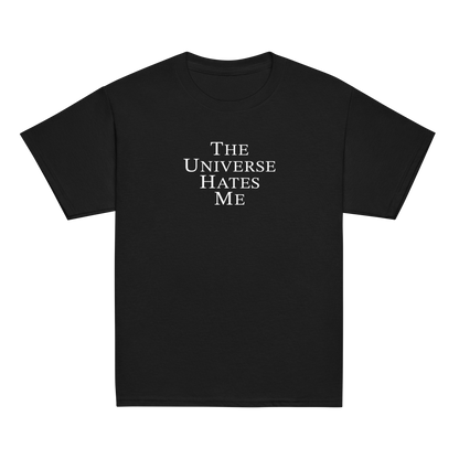 "The Universe Hates Me" printed on a black youth classic tee laying on a flat surface