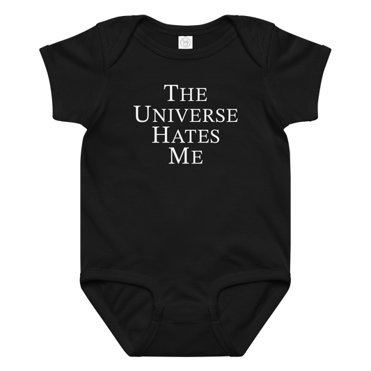 "The Universe Hates Me" printed on a black baby jersey bodysuit laying on a flat surface amongst other clothes