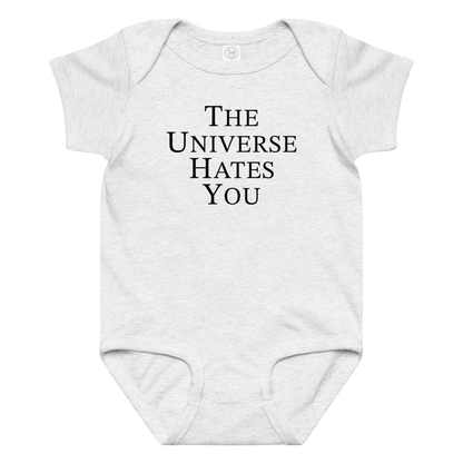 "The Universe Hates You" printed on a ash baby jersey bodysuit laying on a flat surface amongst other clothes