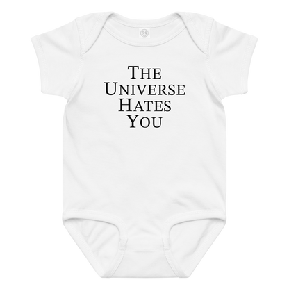 "The Universe Hates You" printed on a white baby jersey bodysuit laying on a flat surface amongst other clothes