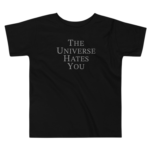 "The Universe Hates You" printed on a black toddler short sleeve tee laying on a flat surface