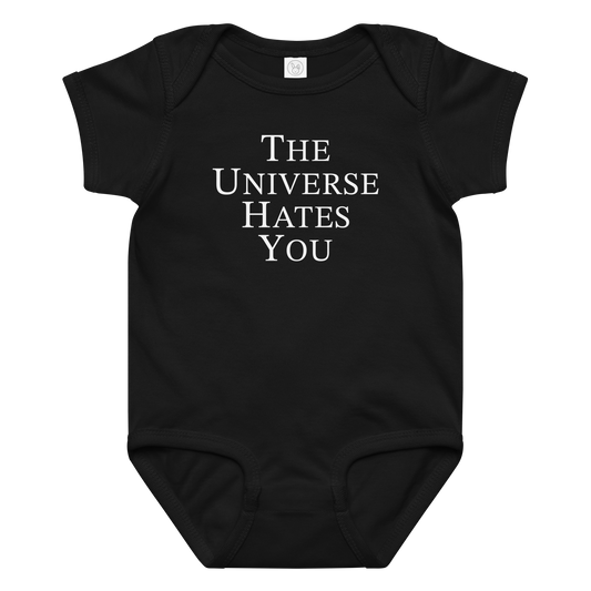 "The Universe Hates You" printed on a black baby jersey bodysuit laying on a flat surface amongst other clothes