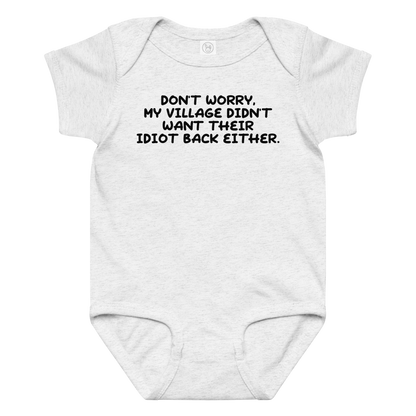 "Don't worry, my village didn’t want their idiot back either." printed on a ash baby jersey bodysuit laying on a flat surface amongst other clothes