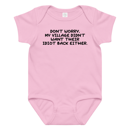 "Don't worry, my village didn’t want their idiot back either." printed on a pink baby jersey bodysuit laying on a flat surface amongst other clothes