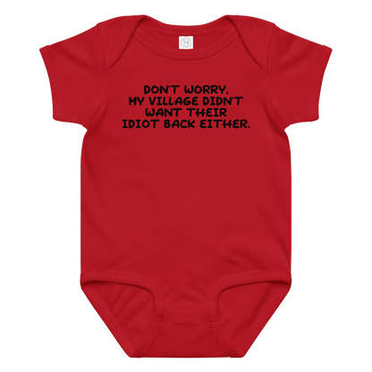 "Don't worry, my village didn’t want their idiot back either." printed on a red baby jersey bodysuit laying on a flat surface amongst other clothes