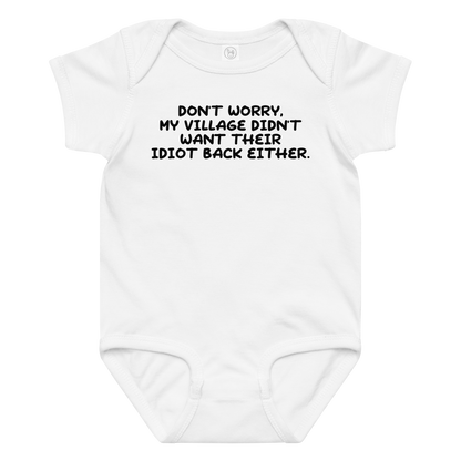 "Don't worry, my village didn’t want their idiot back either." printed on a white baby jersey bodysuit laying on a flat surface amongst other clothes