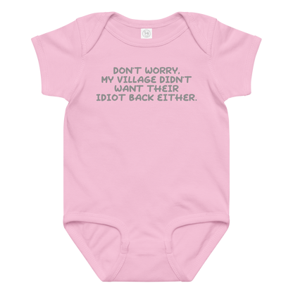 "Don't worry, my village didn’t want their idiot back either." printed on a pink baby jersey bodysuit laying on a flat surface amongst other clothes
