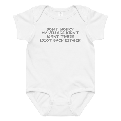 "Don't worry, my village didn’t want their idiot back either." printed on a white baby jersey bodysuit laying on a flat surface amongst other clothes