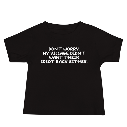 "Don't worry, my village didn’t want their idiot back either." printed on a black baby jersey short sleeve tee laying on a flat surface