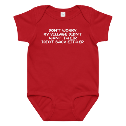 "Don't worry, my village didn’t want their idiot back either." printed on a red baby jersey bodysuit laying on a flat surface amongst other clothes