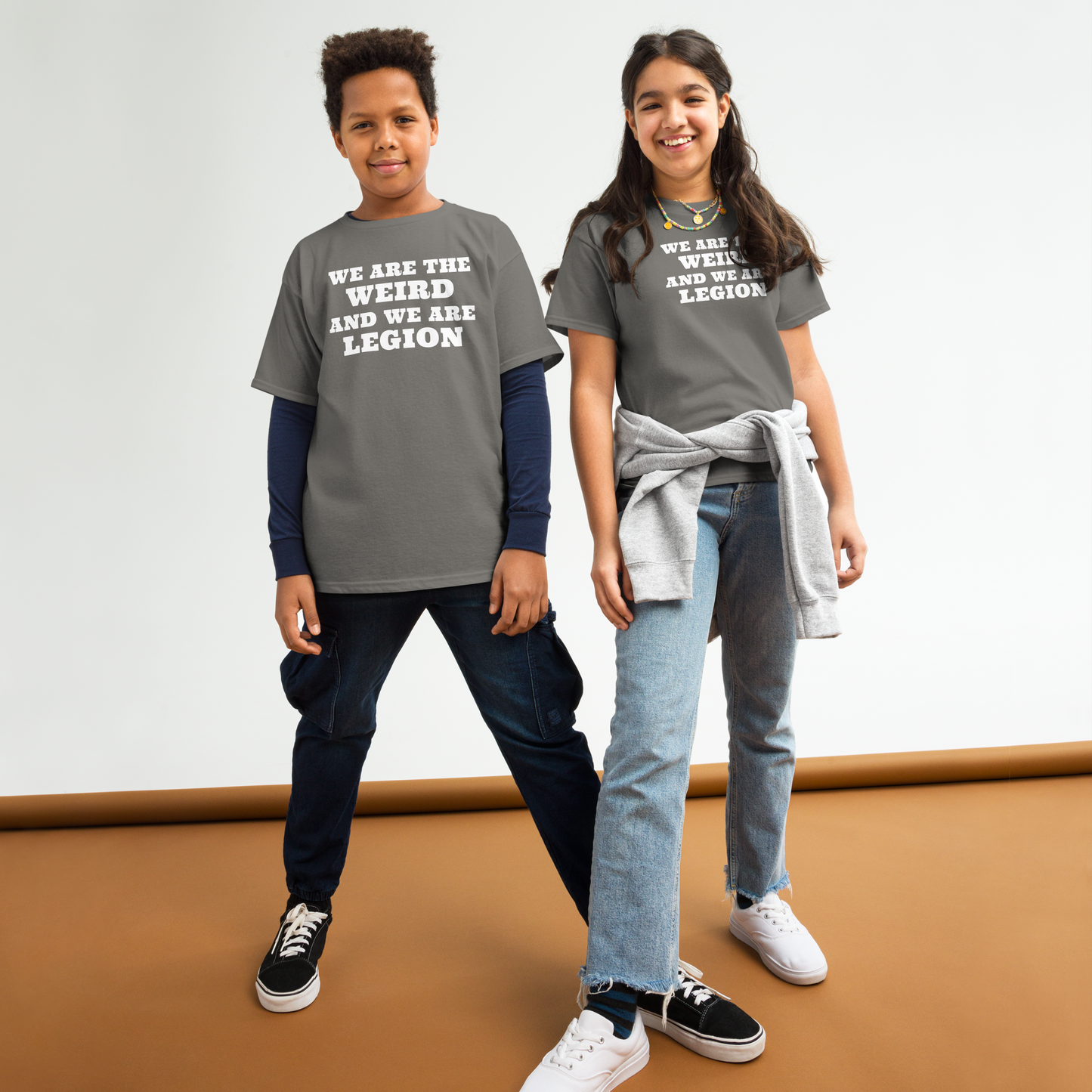 "We are the weird and we are legion" printed on a charcoal youth classic tee worn by a female and male