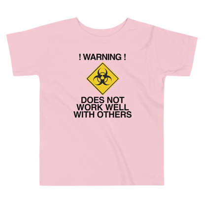 "!Warning! Does Not Work Well With Others" printed on a pink toddler short sleeve tee laying on a flat surface