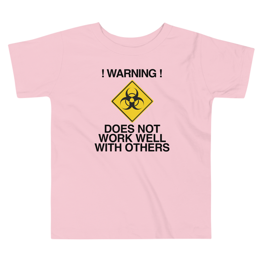 "!Warning! Does Not Work Well With Others" printed on a pink toddler short sleeve tee laying on a flat surface