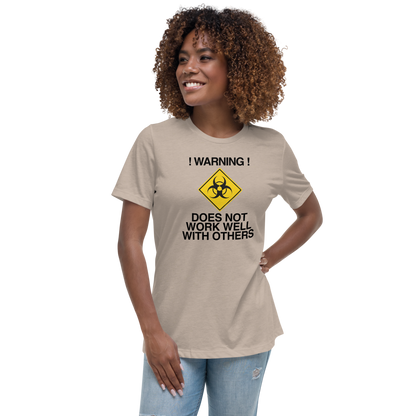 "!Warning! Does Not Work Well With Others" printed on a heather stone women's relaxed t-shirt worn by a female