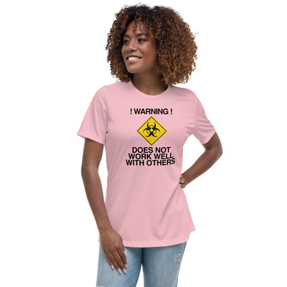 "!Warning! Does Not Work Well With Others" printed on a pink women's relaxed t-shirt worn by a female