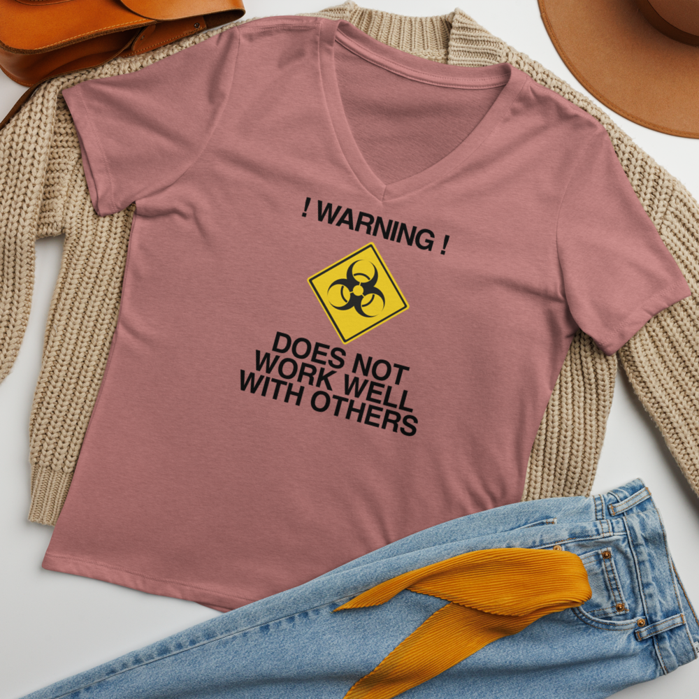 "!Warning! Does Not Work Well With Others" printed on a heather mauve women's relaxed v-neck t-shirt laying on a flat surface amongst other clothes
