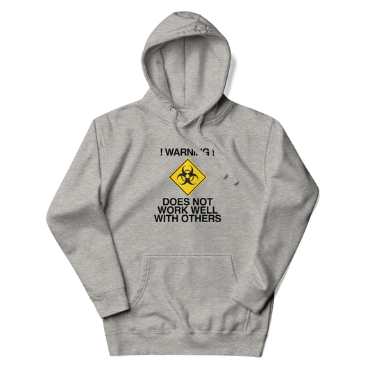 "!Warning! Does Not Work Well With Others" printed on a carbon grey unisex hoodie laying on a flat surface with the hood up
