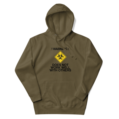 "!Warning! Does Not Work Well With Others" printed on a military green unisex hoodie laying on a flat surface with the hood up