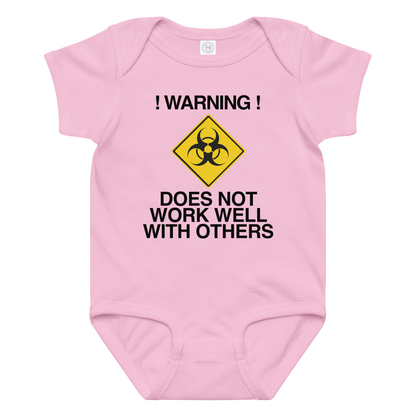 "!Warning! Does Not Work Well With Others" printed on a pink baby jersey bodysuit laying on a flat surface amongst other clothes