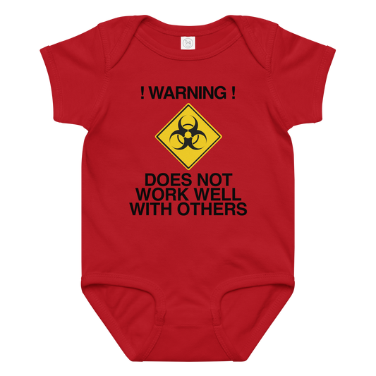 "!Warning! Does Not Work Well With Others" printed on a red baby jersey bodysuit laying on a flat surface amongst other clothes