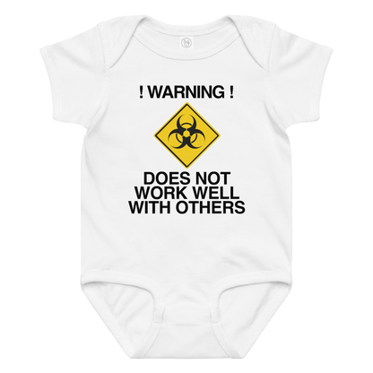 "!Warning! Does Not Work Well With Others" printed on a white baby jersey bodysuit laying on a flat surface amongst other clothes