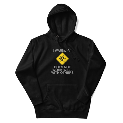 "!Warning! Does Not Work Well With Others" printed on a black unisex hoodie laying on a flat surface with the hood up