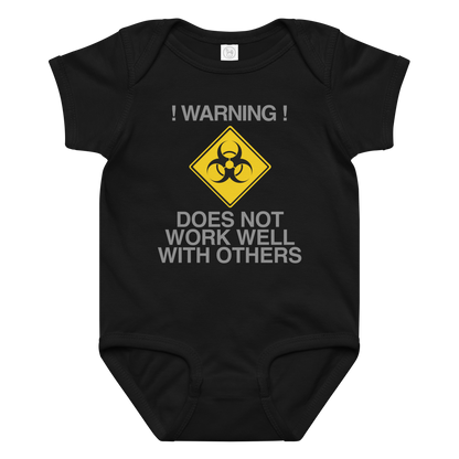 "!Warning! Does Not Work Well With Others" printed on a black baby jersey bodysuit laying on a flat surface amongst other clothes