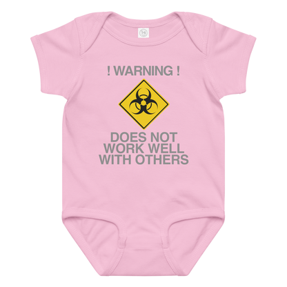 "!Warning! Does Not Work Well With Others" printed on a pink baby jersey bodysuit laying on a flat surface amongst other clothes