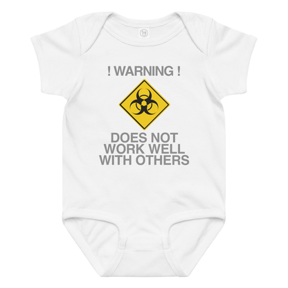 "!Warning! Does Not Work Well With Others" printed on a white baby jersey bodysuit laying on a flat surface amongst other clothes